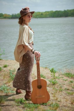 Boho tarzı elbiseleri ve mücevherleri olan güzel bir kızın portresi nehir kıyısında gitarla duruyordu. Yaz tarzı, moda. Hippi kız. Tatil, seyahat. 