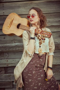 Hippi kız. Boho tarzı kıyafetleri ve mücevherleri olan güzel bir kadın modelin portresi kırsalda eski bir tahta çitin yanında gitarla duruyordu. Yaz stili, tatil. 