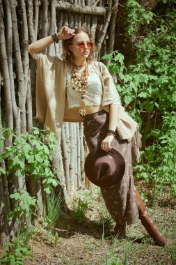 Elbise ve bijouterie giymiş güzel bir kız ahşap bir çitin yanında poz veriyor. Yaz modası. Tatil.