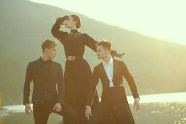 Güzel genç bir bayan birbirine aşık iki yakışıklı adamın arasında duruyor. Deniz kıyısında gün batımında. Aşk üçgeni. 19. yüzyıl tarzı, makyaj, moda..