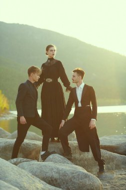 Güzel genç bir bayan birbirine aşık iki yakışıklı adamın arasında duruyor. Deniz kıyısında gün batımında. Aşk üçgeni. 19. yüzyıl tarzı, makyaj, moda..