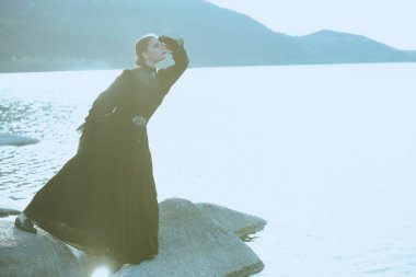 Siyah, zarif bir elbise içinde güzel bir kadın denizin kenarında dikiliyor ve sanki birini bekliyormuş gibi dikkatlice ve gergin bir şekilde uzaklığa bakıyor. 19. yüzyıl romanı. Boşluğu kopyala.