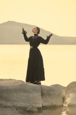 Siyah zarif elbiseli genç bir kadın deniz kenarında üzgün bir şekilde duruyor. Duygularından muzdarip ve merakla gökyüzüne bakıyor. 19. yüzyıl romanı. 