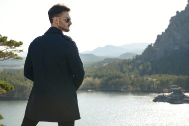 Klasik takım elbiseli ve siyah güneş gözlüklü kendine güvenen bir iş adamı dağın zirvesinde durmuş güzel doğaya bakıyor. İş, başarı. Tatil ve seyahat, iş gezisi. Boşluğu kopyala.