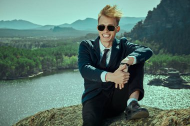 Klasik takım elbiseli ve siyah güneş gözlüklü yakışıklı genç bir adam bir dağ manzarası ve neşeyle gülümseyen bir göle karşı oturur. İş, başarı. Tatil ve seyahat, iş gezisi..