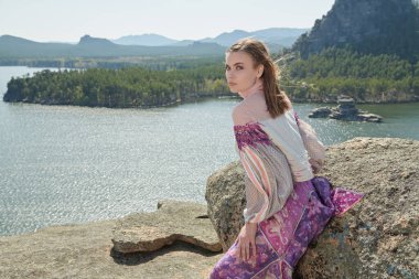 Güzel bir gölün arka planındaki bir dağda duran şık etnik kıyafetlerle poz veren güzel bir kız modeli. Moda ve etnik tarz. Seyahat ve tatil. 