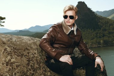 Deri ceketli, siyah güneş gözlüklü ciddi bir sarışın manken deniz kıyısının zeminine karşı dağlarda poz veriyor. Erkek modası ve deri giyim tarzı. 