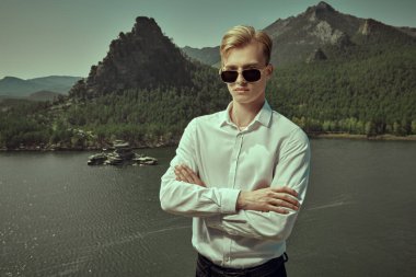 Beyaz gömlekli ve siyah güneş gözlüklü kasıtlı genç bir adam kollarını çapraz bir şekilde bir göle ve dağ manzarasına dayar. İş konsepti. Tatil ve seyahat.