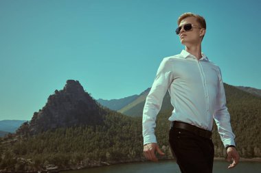 Beyaz gömlekli, siyah güneş gözlüklü yakışıklı ve kasıtlı bir adam bir dağ manzarası ve göle karşı duruyor. İş konsepti, başarı. Tatil ve seyahat. Boşluğu kopyala.