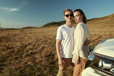 Sevgililer tatilde. Yakışıklı bir genç adam ve güzel bir kız gün batımında bozkırda arabalarının yanında birbirlerine sarılıyorlar. Yaz stili. Tatil, yaz tatili ve seyahat. Boşluğu kopyala.