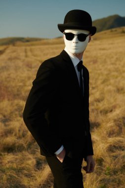 Siyah takım elbiseli, melon şapkalı ve beyaz maskeli gerçek üstü bir beyefendi vadinin arka planında dikiliyor..