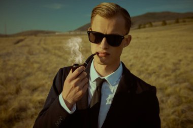 Siyah takım elbiseli ve siyah güneş gözlüklü zarif bir beyefendi vadide dikilip pipo içiyor..