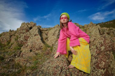 Kıyafetler ve aksesuarlar içinde parlak bir yaz stili. Şık genç kadın parlak pembe ve yeşil giysiler ve pembe güneş gözlükleriyle bir dağın tepesinde mavi gökyüzüne karşı poz veriyor. Yüksek moda koleksiyonu.