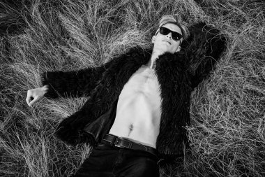 Çıplak vücudunun üzerinde siyah kürk giyen yakışıklı bir mankenin siyah beyaz portresi ve bozkırdaki kuru çimlerin üzerinde yatan siyah kot. Yüksek moda çekim. 