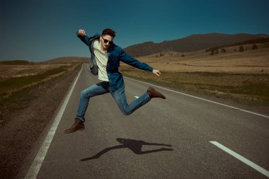Sıradan kotlar giyen çekici genç bir adam otoyolda neşeyle zıplıyor. Denim modası. Yol maceraları, seyahat, otostop.. 