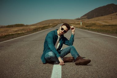 Sıradan kotlar giyen çekici genç bir adam otobanda oturmuş geçen bir arabayı bekliyor. Yol seyahati otostop yapıyor. Denim modası. 