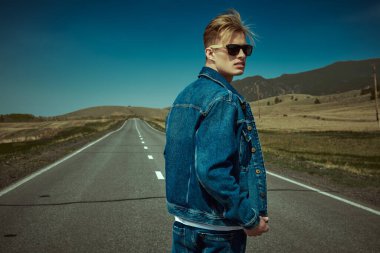 Kot elbiseli, siyah güneş gözlüklü yakışıklı otostopçu resimli bir manzaranın üzerinde duruyor. Denim modası. Yol maceraları, seyahat.