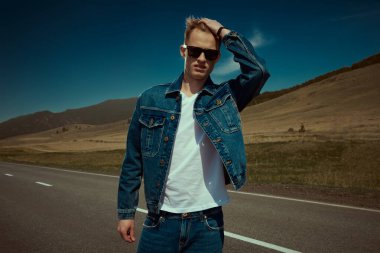 Kot pantolon ve siyah güneş gözlüklü çekici bir otostopçu manzaranın arka planında duruyor. Denim modası. Yol maceraları, seyahat.