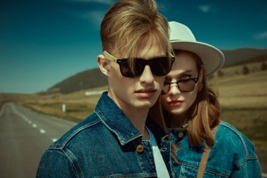 Tepe vadisindeki bir otoyolda duran kot elbiseli ve siyah güneş gözlüklü güzel bir çiftin portresi. Denim modası. Yol seyahati otostop yapıyor. Özgürlük.