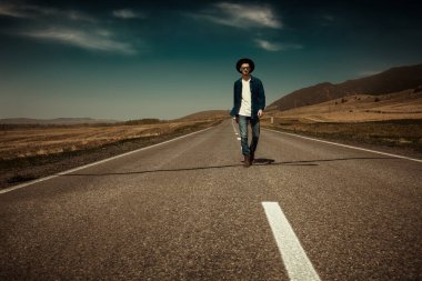 Şık kot elbiseli ve güneş gözlüklü yalnız bir gezgin vadide koşan bir otoyolda yürüyor. Yol seyahati otostop yapıyor. Denim modası. Boşluğu kopyala.