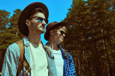 İki yakışıklı sırt çantalı adam bir çam ormanının arka planında yan yana durur. Hippiler dışarıda. Yürüyüş, seyahat ve turizm.