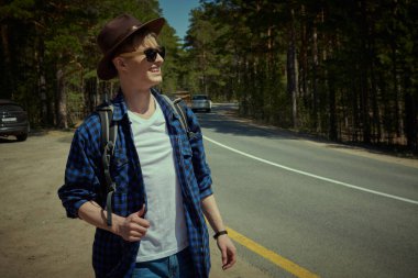 Mutlu, genç bir sırt çantalı adam çam ormanının arka planında neşeli bir gülümsemeyle dikiliyor. Hipster açık havada. Otostop, seyahat ve turizm.
