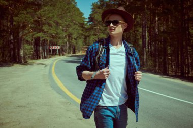 Yakışıklı bir sırt çantalı adamın portresi şık günlük giysiler içinde bir çam ormanının arka planında dikilip başka yöne bakıyor. Hipster açık havada. Yürüyüş, seyahat ve turizm.
