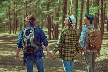 Mutlu genç insanlar sırt çantalı arkadaşlar çam ormanında el ele tutuşarak yol boyunca yürürler. Yaz turizmi, açık hava aktiviteleri, yürüyüş..