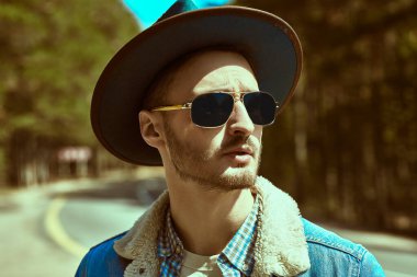 Kot ceketli, yakışıklı, sakallı bir adamın portresi, otobanın kenarında duran siyah bir şapka ve güneş gözlüğü. Denim modası. Yol maceraları, otostop, otostop.. 
