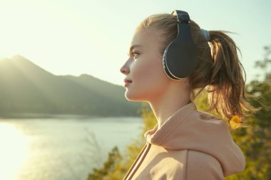 Mutlu, gülümseyen bir kız gün batımında güzel bir deniz ve dağ manzarasına karşı kulaklıkla hoş bir müzik dinler. Seyahat, yaz tatilleri. Doğayla uyum..