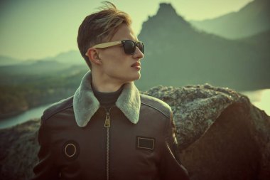 Deri ceketli ve siyah güneş gözlüklü yakışıklı genç bir adam dağın tepesinde duran mesafeye bakıyor. Dağ manzarası arka planı. 