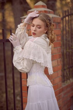 Viktorya döneminin tarihi yeniden yapılandırması. Beyaz elbiseli güzel bir hanımın portresi saray çitinde duruyor. Saç ve makyaj, 19. yüzyılın sonlarında..