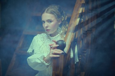 Tarihsel mistik roman. Beyaz dantel elbiseli güzel bir bayan aristokrat, kameraya şüpheyle bakan puslu, karanlık bir vintage odadaki ahşap merdivenlerde oturuyor. Viktorya tarzı.