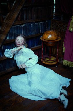 Beyaz dantelli elbiseli hoş bir aristokrat bayan karanlık bir kütüphanede kitaplığın yanında oturuyor. Viktorya tarzı. Tarihi roman..