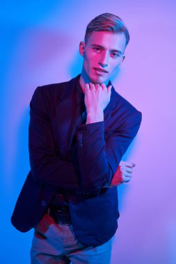 Şık gömlek, ceket ve pantolon giymiş çekici, genç bir sarışının moda portresi. Duvarın yanında çok renkli bir ışıkla duruyor. Erkek modası. 