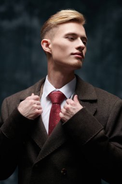 Çekici sarışın bir adam pahalı ve zarif bir ceket, beyaz gömlek ve koyu bir arka planda kravat ile poz veriyor. Erkek güzelliği, moda. 