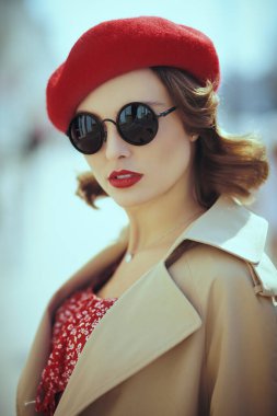 Zarif bir retro tarzı. Kırmızı elbiseli, yağmurluklu, kırmızı bereli ve zarif siyah güneş gözlüklü güzel bir kadın bir şehir caddesinde duruyor. Güzellik, moda.
