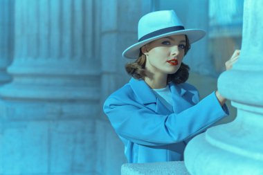 Zarif bir gökyüzü mavi takım elbiseli, beyaz şapkalı güzel bir esmer kadının portresi. Şehrin caddesinde binanın bir köşesinde duruyor. 40 'lı yılların tarzı. Makyaj ve saç stili. Hollywood tarzı. 