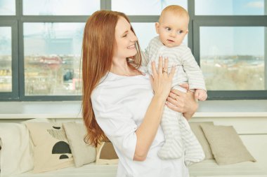 Mutlu, gülümseyen bir anne, bir apartmanın penceresinde duran küçük bebeğine şefkatle bakar. Mutlu annelik.