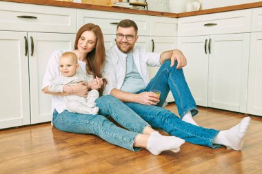 Mutlu aile fotoğrafı. Neşeli anne ve baba, sevimli bebekleriyle mutfak zemininde oturuyor. Modern iç mekan, mutfak mobilyası.. 