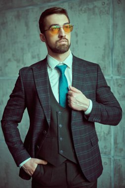 Zarif takım elbiseli ve güneş gözlüklü kendinden emin orta yaşlı bir işadamı gri bir duvar arkasında poz veriyor. Erkek modası.