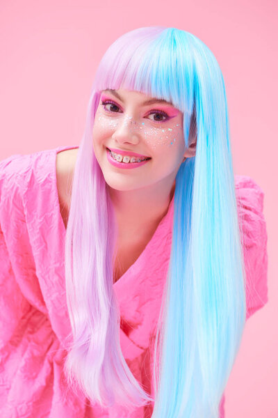 A smiling girl with bright makeup and in colored violet-blue wig posing in stylish pink dress. Pink background. Fashion. Hairstyle, hair coloring, make-up. Japanese anime style. 