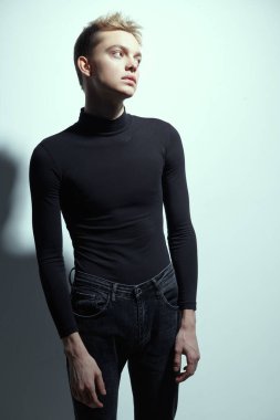 Siyah balıkçı yaka kazak ve kot pantolon giymiş beyaz bir duvara poz veren erkek mankenin sakin ve düşünceli bir şekilde çekilmiş bir fotoğrafı. Moda fotoğrafı.. 