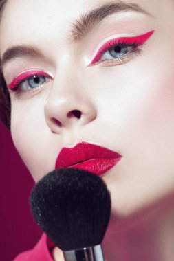 Parlak pembe rujlu ve gözlerinde pembe oklar olan güzel, sarışın bir kız yüzüne yakın bir makyaj fırçası tutuyor. Fuchsia geçmişi. Makyaj ve makyaj malzemeleri. Makyaj araçları.