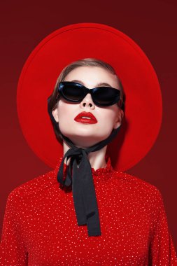 Parlak makyajlı, zarif kırmızı elbise, kırmızı şapka ve güneş gözlüğü takan güzel bir kadın. Burgonya arkaplanı. Moda fotoğrafı. Makyaj ve kozmetik. 