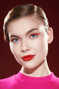 Kozmetik ve makyaj malzemeleri. Parlak kırmızı rujlu ve gözlerinde kırmızı oklar olan sevimli sarışın bir kız kameraya hafifçe gülümsüyor. Burgonya arkaplanı. Güzellik konsepti.