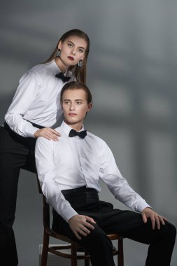 Beyaz gömlekli, papyonlu ve siyah pantolonlu güzel kadın ve yakışıklı genç adam, ışıktan gölgeli gri bir arka plana karşı poz veriyorlar. İş stili. Başarılı insanlarız. Moda çekimi.. 