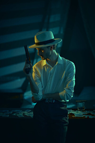 A pensive man with a gun in his hand stands leaning on a table with banknotes and playing cards in the bedroom with dark blue lighting. Retro style. Mafia, gambling and the criminal world.