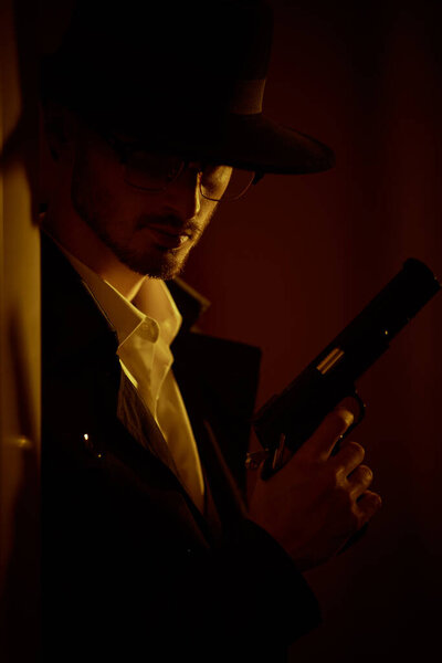 Criminal world, mafia. Portrait of a thoughtful brunet man in black coat and a hat standing in dark room, lit by a ray of light, with a gun in his hand. Retro noir style. 