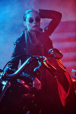 Zarif bir gece elbisesi, siyah deri ceketi ve garajda motosikletiyle şık bir güneş gözlüğü takan güzel bir motosikletli kız..